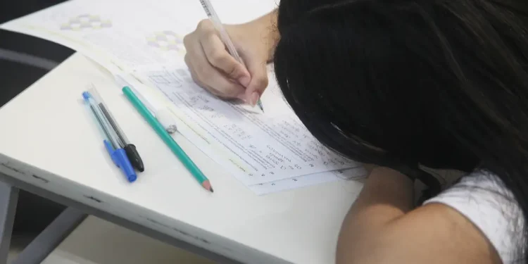 Jovem estudante concentrada escreve em folha de prova sobre carteira escolar em imagem ilustrativa sobre as novas regras do Enem 2026