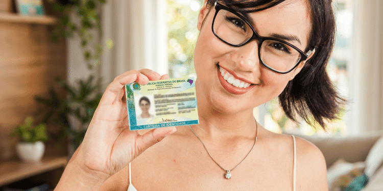 Mulher de óculos sorri e segura a Carteira de Identidade Nacional próxima ao rosto.