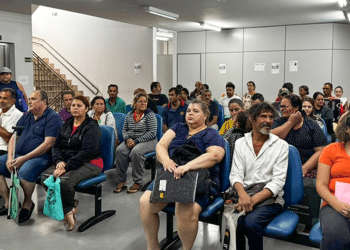 Pessoas aguardam atendimento sentadas em sala de espera de agência do INSS, perto do setor de perícia médica.