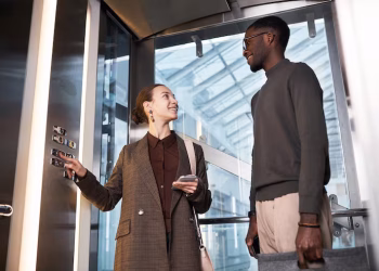 Duas pessoas conversando dentro de um elevador de vidro em prédio moderno
