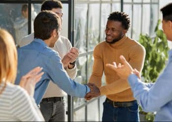 Colegas sorriem e se cumprimentam e, ao redor, o corredor claro de um ambiente