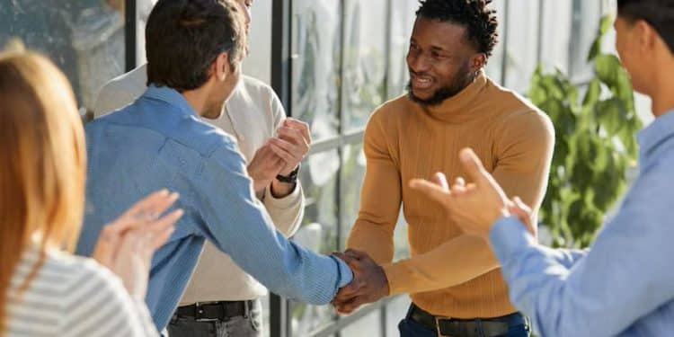Colegas sorriem e se cumprimentam e, ao redor, o corredor claro de um ambiente