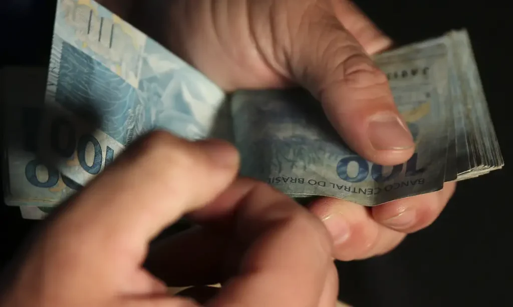 Mãos manuseando notas de cem reais durante recebimento de valores acima do salário mínimo do INSS