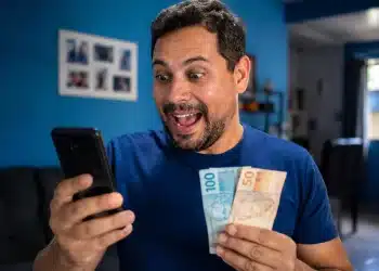 Homem sorridente consultando aplicativos no celular segurando notas de dinheiro, representando a liberação do pagamento de R$ 3.981.