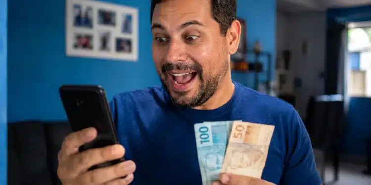 Homem sorridente consultando aplicativos no celular segurando notas de dinheiro, representando a liberação do pagamento de R$ 3.981.