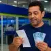 Brasileiro consulta comprovante de pagamento e segura notas de R$ 100 em agência bancária, representando o pagamento de R$ 978,01.