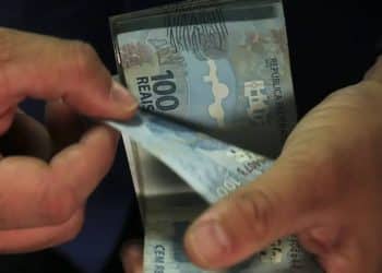Mãos contando notas de 100 reais com destaque para cédulas azuis e dedos masculinos, simbolizando o pagamento de R$ 1.216 do Abono Salarial PIS/Pasep para quem trabalhou 9 meses no ano-base.