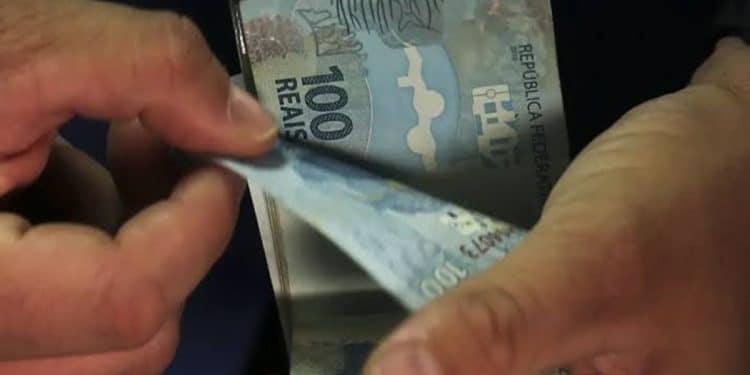 Mãos contando notas de 100 reais com destaque para cédulas azuis e dedos masculinos, simbolizando o pagamento de R$ 1.216 do Abono Salarial PIS/Pasep para quem trabalhou 9 meses no ano-base.