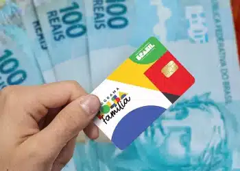 Mão segura cartão do Programa Bolsa Família com chip dourado sobre várias notas de R$100 azuis ao fundo, simbolizando o extra de R$ 300.
