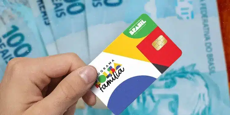 Mão segura cartão do Programa Bolsa Família com chip dourado sobre várias notas de R$100 azuis ao fundo, simbolizando o extra de R$ 300.