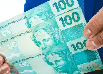 Notas de 100 reais sendo seguradas nas mãos, representando pagamento de R$ 127 a R$ 1.518 liberado para CPFs de finais 0 a 9.
