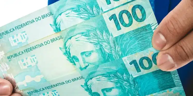 Notas de 100 reais sendo seguradas nas mãos, representando pagamento de R$ 127 a R$ 1.518 liberado para CPFs de finais 0 a 9.