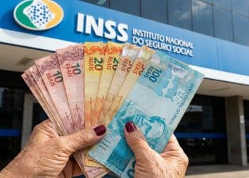Aposentada exibe dinheiro em mãos diante da fachada azul de uma agência do INSS em dia ensolarado.