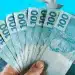 Mão segurando notas de R$ 100 representando o pagamento de R$ 4.800 ou mais para titulares de CPFs com finais de 0 a 9.