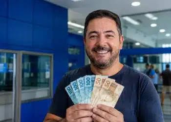 Homem segurando notas de R$ 100 e R$ 50 em agência bancária, representando pagamento liberado de até R$ 6.220 para CPFs com finais de 0 a 9.