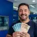 Homem segurando notas de R$ 100 e R$ 50 em agência bancária, representando pagamento liberado de até R$ 6.220 para CPFs com finais de 0 a 9.