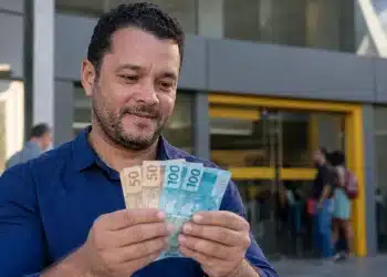 Homem segurando cédulas de real em frente a uma agência bancária, representando o saque do PIS/Pasep de junho.