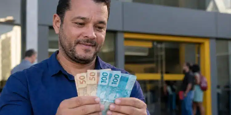 Homem segurando cédulas de real em frente a uma agência bancária, representando o saque do PIS/Pasep de junho.