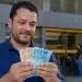 Homem segurando cédulas de real em frente a uma agência bancária, representando o saque do PIS/Pasep de junho.