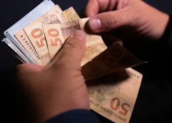 Mãos contam várias cédulas de cinquenta reais sobre fundo escuro.