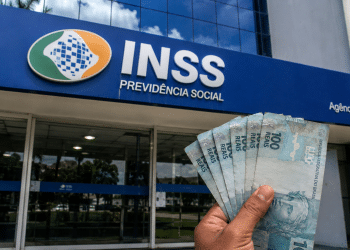 Mão segurando cédulas de cem reais em frente a uma agência do INSS, em referência ao pagamento de R$ 1.621 ou R$ 2.431,50 da segunda parcela do 13º salário em 2026.
