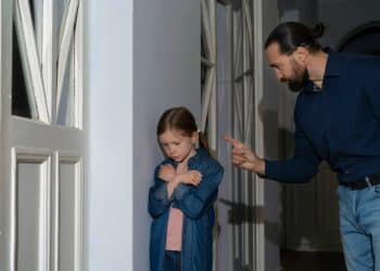 Homem adulto apontando dedo para menina ruiva de braços cruzados em postura defensiva, em corredor residencial, simbolizando o narcisismo parental.
