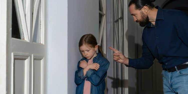 Homem adulto apontando dedo para menina ruiva de braços cruzados em postura defensiva, em corredor residencial, simbolizando o narcisismo parental.