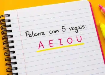 Caderno espiral aberto com a frase “Palavra com 5 vogais: A E I O U” escrita em destaque colorido sobre fundo amarelo vibrante.