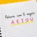 Caderno espiral aberto com a frase “Palavra com 5 vogais: A E I O U” escrita em destaque colorido sobre fundo amarelo vibrante.