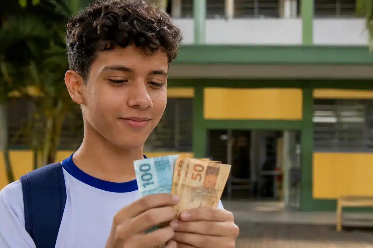 Estudante do ensino médio segurando dinheiro em frente à escola, representando os R$ 200 extras do Pé-de-Meia e a isenção no Enem 2026 para concluintes.