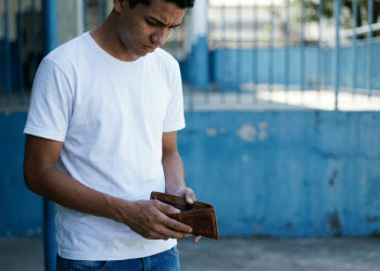 Jovem de camiseta branca olha preocupado para uma carteira aberta e vazia em um pátio escolar