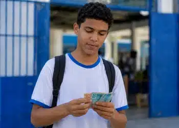 Estudante segurando notas de real na entrada de escola pública, representando os últimos a receber os pagamentos do Pé-de-Meia de maio.