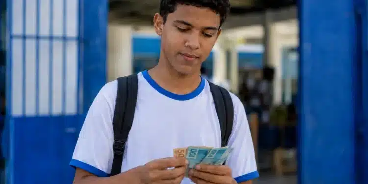 Estudante segurando notas de real na entrada de escola pública, representando os últimos a receber os pagamentos do Pé-de-Meia de maio.