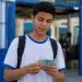 Estudante segurando notas de real na entrada de escola pública, representando os últimos a receber os pagamentos do Pé-de-Meia de maio.