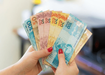 Mãos com unhas vermelhas exibem notas de 50, 10, 20 e 100 reais em formato de leque, mostrando a segunda família do real, simbolizando o pagamento de R$ 4.116.