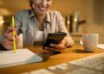 Mulher sorridente segura o celular em uma das mãos e um lápis na outra diante de caderno aberto e teclado sobre mesa de madeira