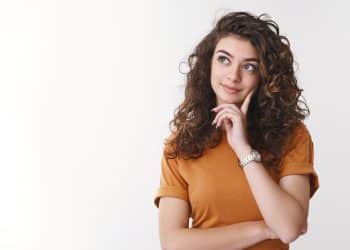 Jovem mulher de cabelo cacheado e camiseta laranja com a mão no rosto em expressão pensativa sobre fundo branco