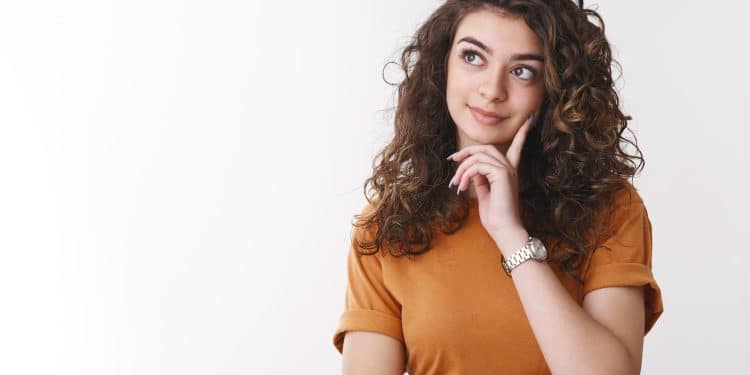 Jovem mulher de cabelo cacheado e camiseta laranja com a mão no rosto em expressão pensativa sobre fundo branco