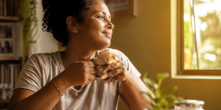 Mulher segura uma xícara e sorri olhando pela janela em um ambiente tranquilo de casa