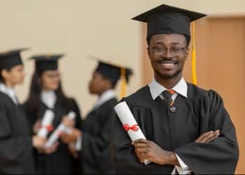 Jovem formando de beca e capelo sorri segurando o diploma