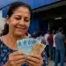 Mulher segurando cédulas de real em frente a banco, simbolizando prazo final para solicitar pagamento de R$ 2,9 mil.