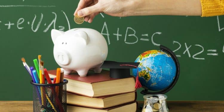Mão com blusa amarela colocando moeda em cofrinho porquinho branco sobre livros, ao lado de globo e lousa verde, simbolizando as possibilidades de renda extra para formados em pedagogia.