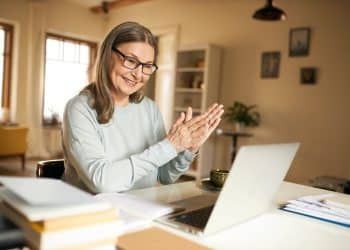 Mulher de cabelos castanhos e óculos aplaude sorridente diante de laptop em escritório doméstico iluminado, simbolizando o salário de um professor EAD no Brasil.