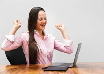 Mulher de camisa rosa comemora em frente ao notebook, simbolizando as profissões com salários de R$ 7 mil.