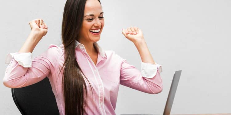 Mulher de camisa rosa comemora em frente ao notebook, simbolizando as profissões com salários de R$ 7 mil.