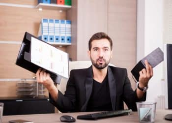 Homem de barba com blazer preto segura prancheta com gráficos na mão direita e tablet na esquerda, com expressão de surpresa, simbolizando as profissões em que faltam profissionais.