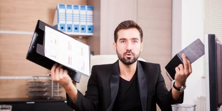 Homem de barba com blazer preto segura prancheta com gráficos na mão direita e tablet na esquerda, com expressão de surpresa, simbolizando as profissões em que faltam profissionais.