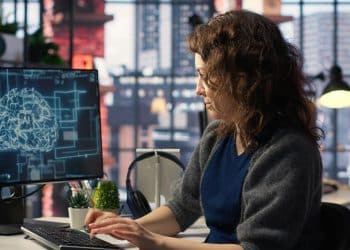 Mulher de cabelos cacheados digita em teclado observando cérebro holográfico com sigla AI na tela do monitor, simbolizando as profissões que mais podem crescer com a inteligência artificial.