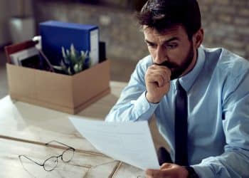 Homem preocupado analisando documento após perda de emprego em escritório.
