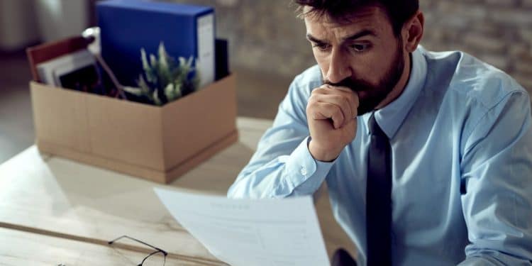 Homem preocupado analisando documento após perda de emprego em escritório.